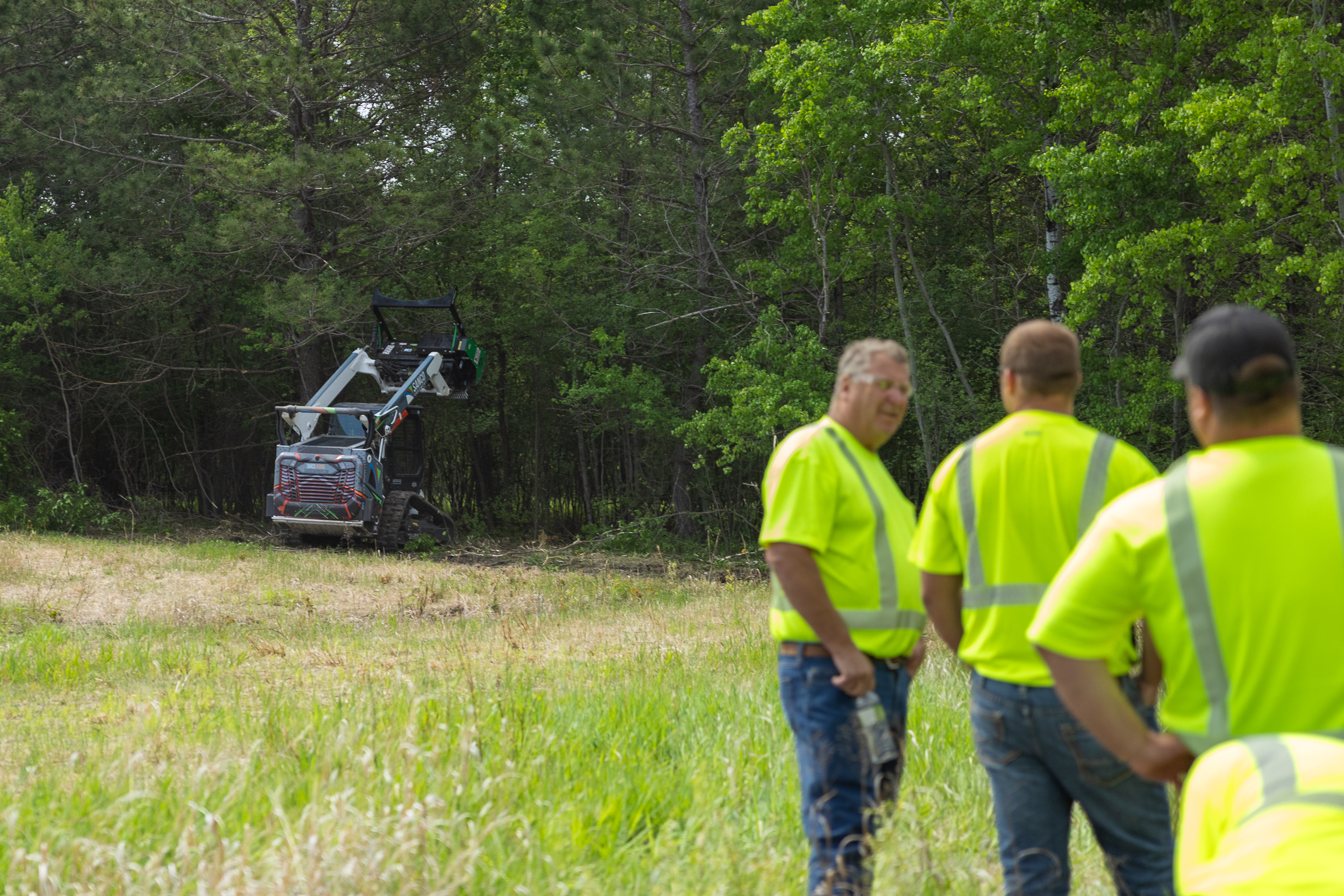 people standing around talking while a skid steer drum mulcher mulches trees in the background